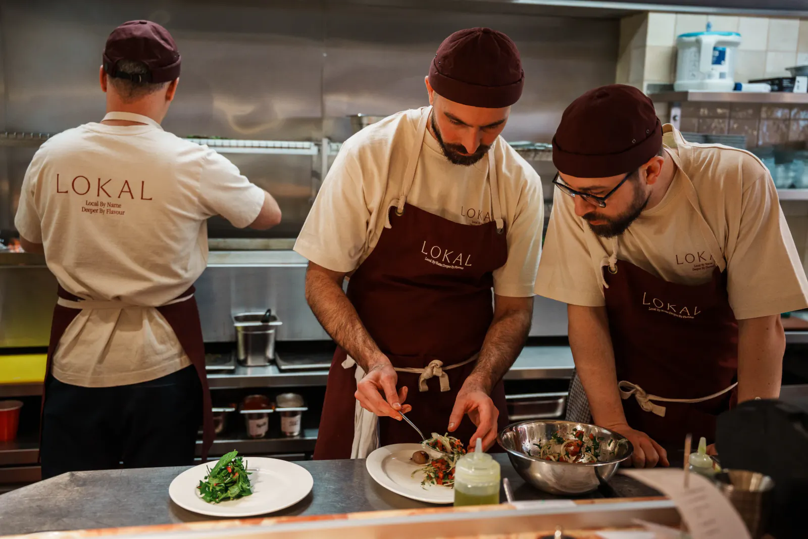 Three chefs in LOKAL branded uniforms plating food in a professional kitchen