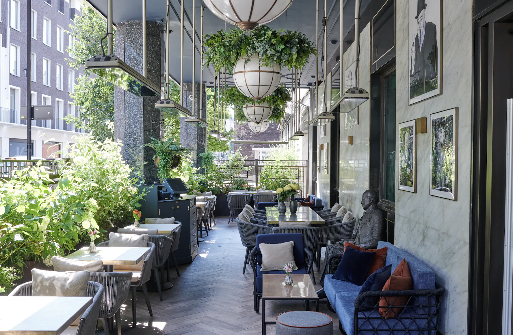 Modern outdoor restaurant patio with blue sofa seating, hanging plants, framed art on marble walls, and city buildings visible