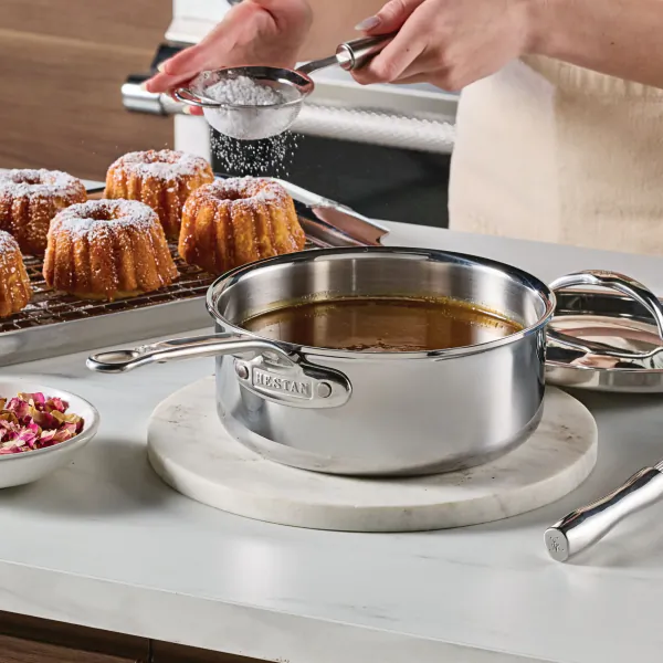 Person dusting powdered sugar over Bundt cakes with a sieve, stainless steel Hestan saucepan with caramel sauce on marble trivet in foreground