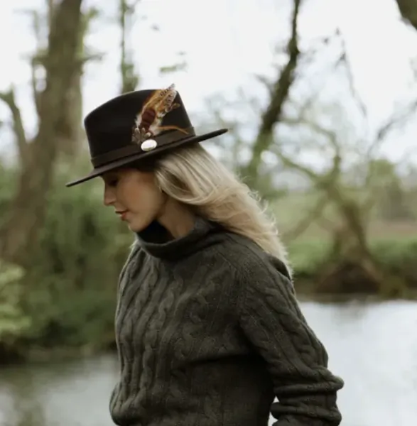 Woman in profile wearing a dark wide-brim hat with feathers and a chunky knit sweater, standing by a misty river in a wooded setting