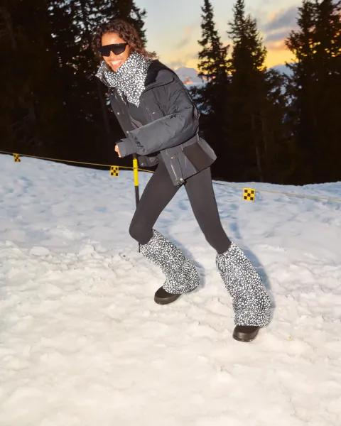 Woman in grey puffer jacket and leopard-print leg warmers posing on snowy mountain slope at sunset