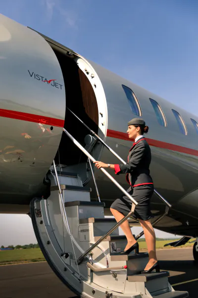 Flight attendant in dark uniform descending the stairs of a silver VistaJet private plane on the tarmac