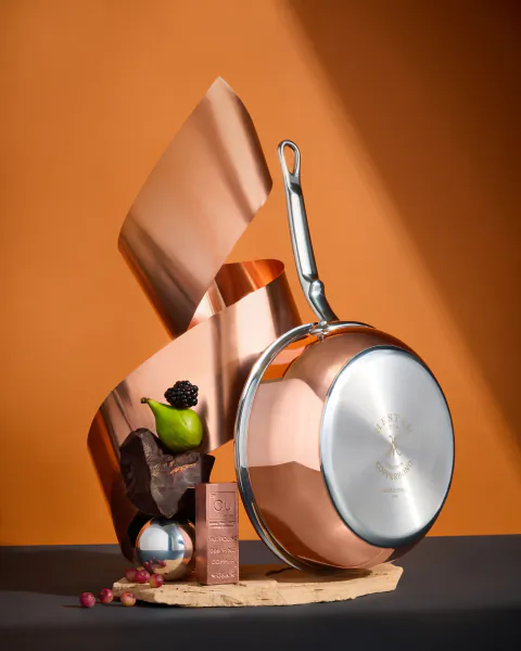 Hestan Culinary copper frying pan with silver handle on wooden board, surrounded by chocolate bar, blackberry, green pear, and copper sheet against orange background