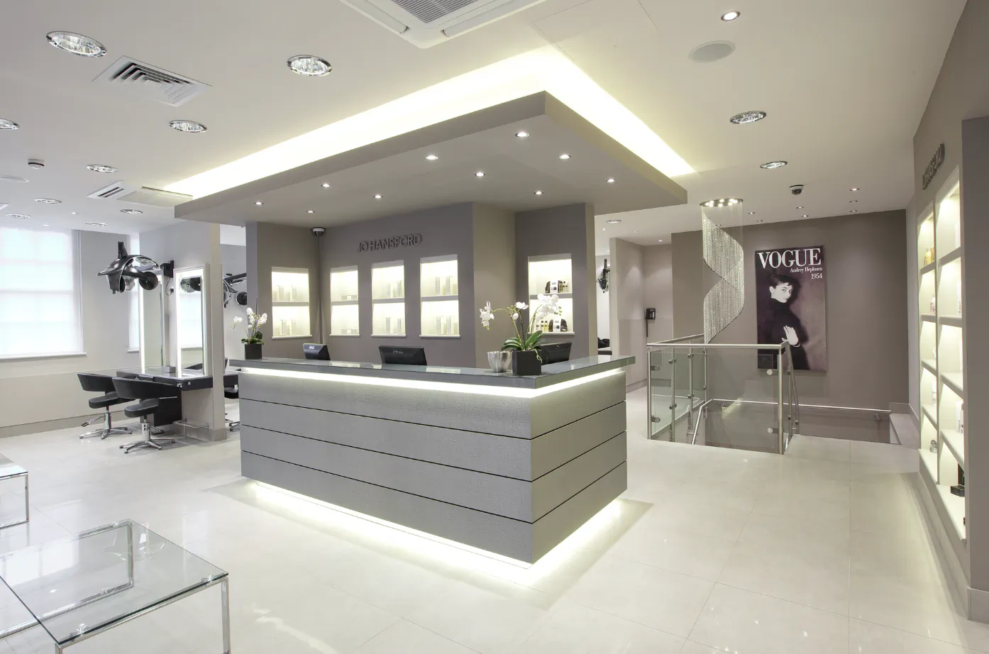 Modern luxury hair salon reception with illuminated grey desk, 'Jo Hansford' signage, Vogue poster, and styling stations