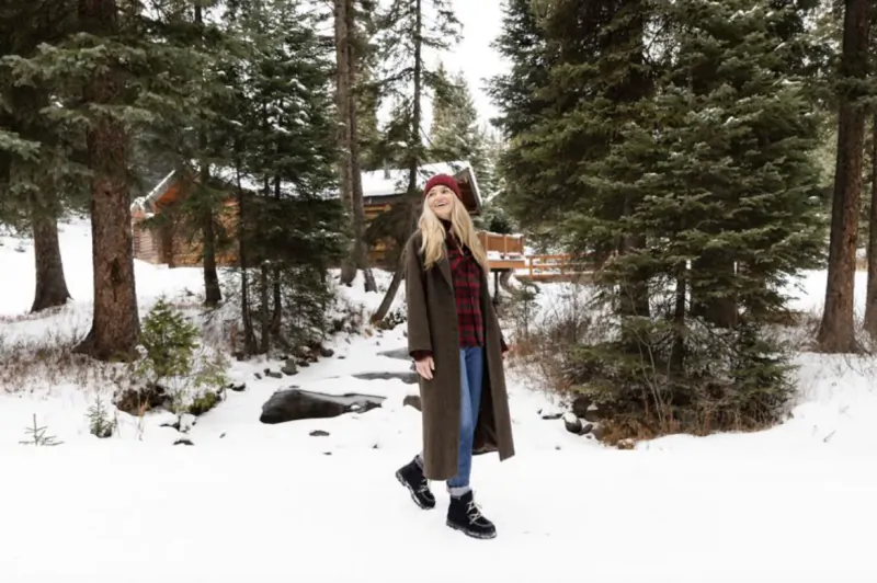 Woman in red beanie and long coat standing in snowy forest path near wooden cabin