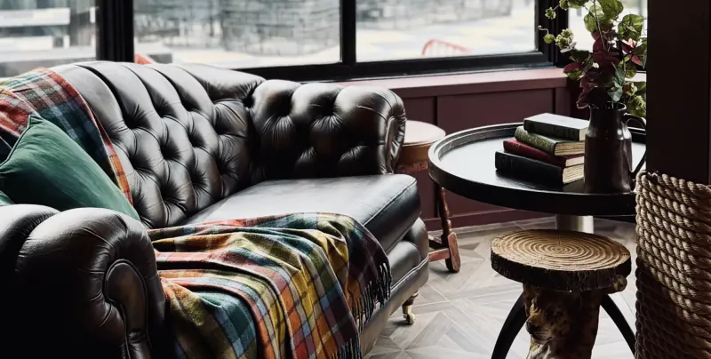 Black tufted leather Chesterfield sofa with colorful plaid throw and green cushion beside round table with stacked books and vase in modern living room