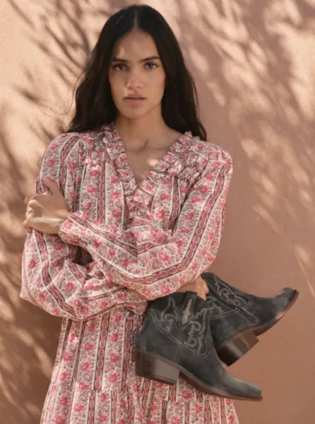 Woman in floral print dress holding grey embroidered cowboy boots against a pink wall